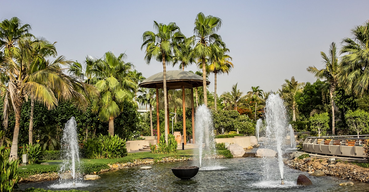 Jardim de palmeiras com lago, fontes de água e estrutura central para descanso