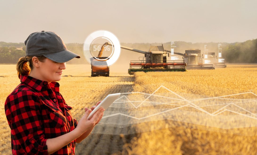 Mulher vestindo camisa xadrez segurando tablet em meio ao campo. Ao fundo, colhedoras trabalhando na colheita de grãos com caminhão transbordo ao lado.