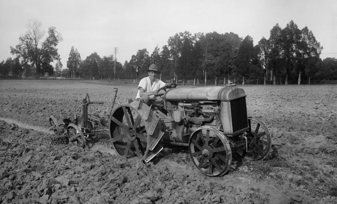 Imagem em preto e branco mostrando um homem em um dos primeiros modelos de tratores movidos a combustão. O trator conta com rodas de ferro e implemento em campo.