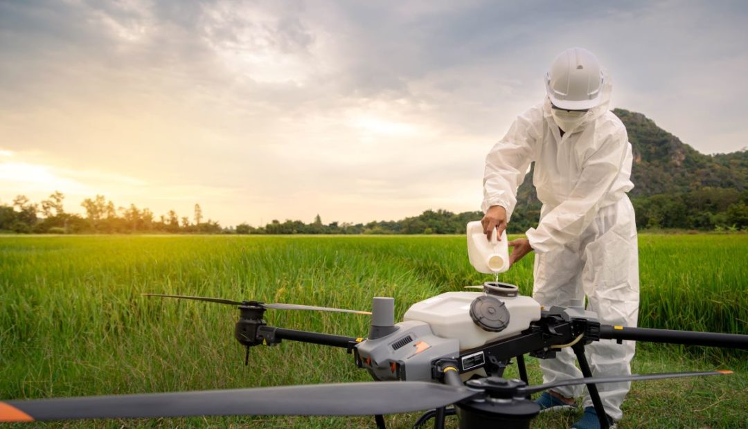 Pessoa utilizando equipamento de proteção individual enquanto faz o abastecimento de drone em meio a plantação, visando a segurança operacional.