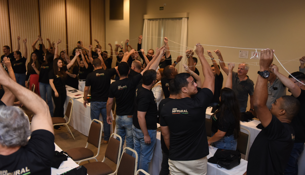 Dinâmica entre os colaboradores durante o evento. Colaboradores vestem camiseta preta e seguram barbantes entrelaçados no alto.