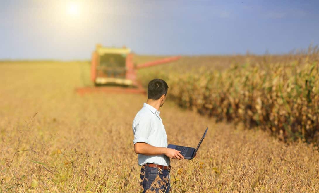 Homem com laptop em mãos, olhando para colhedora de grãos em campo.