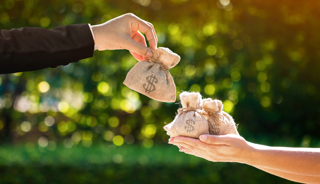 Mão de homem com terno entregando saco de dinheiro para mãos femininas estendidas já com outros sacos de dinheiro. Ao fundo, vegetação.