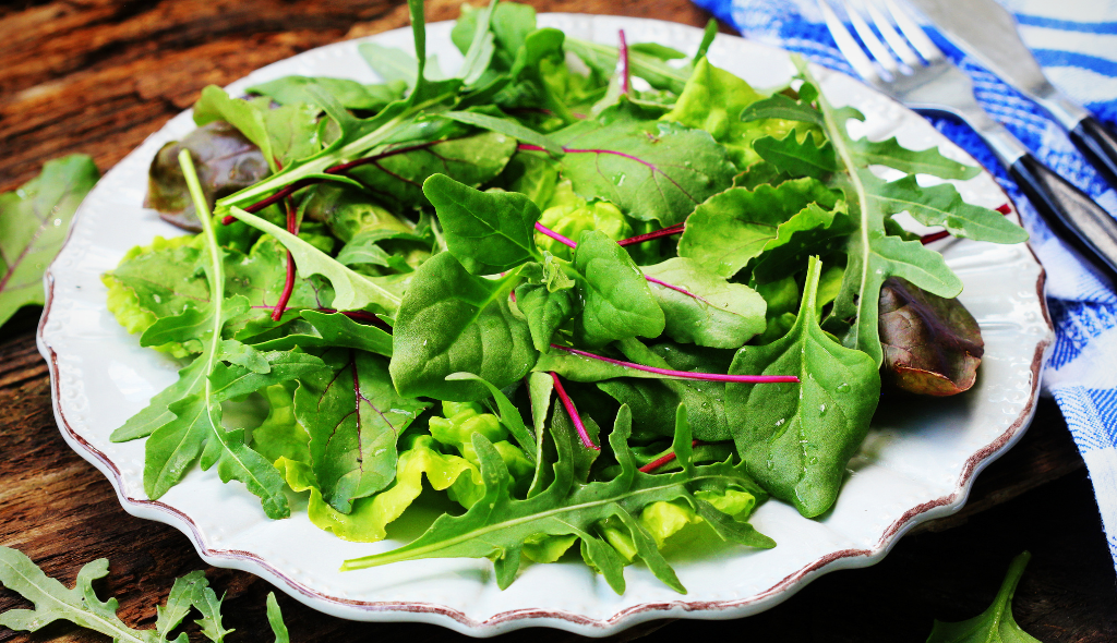 Prato de salada contendo folhas de espinafre, ao lado do prato há um pano de prato branco e azul, com garfo e faca em cima