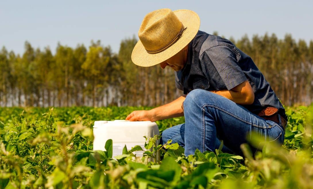 Engenheiro agrônomo agachado em campo de soja com galão de defensivo agrícola em mãos