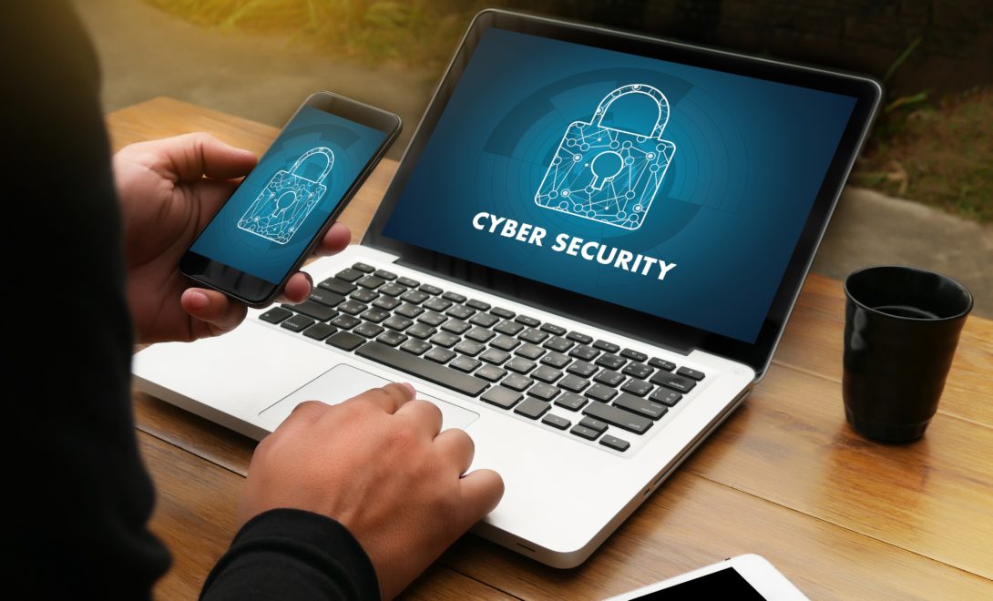 Homem segurando smartphone manuseando um laptop, ambos com imagens de cadeado escrito cyber security (segurança cibernética)