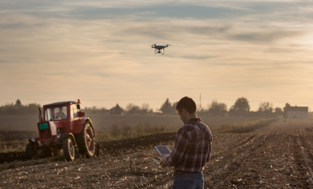 Homem segurando controlador de um drone que está sobrevoando uma área observando colheita feira por trator