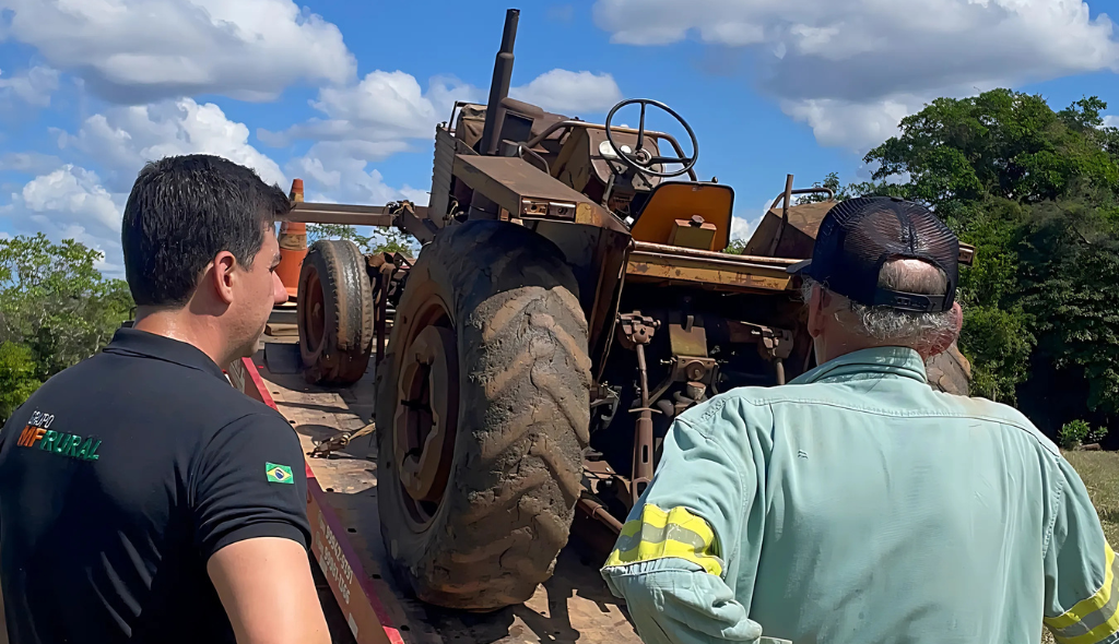 Rafael Fabrizzi Lucas, CEO do MF Rural, e Seu Clarismundo observam juntos o trator Valmet 110 de 1974 carregado em um caminhão prancha, em meio a um dia ensolarado no campo.
