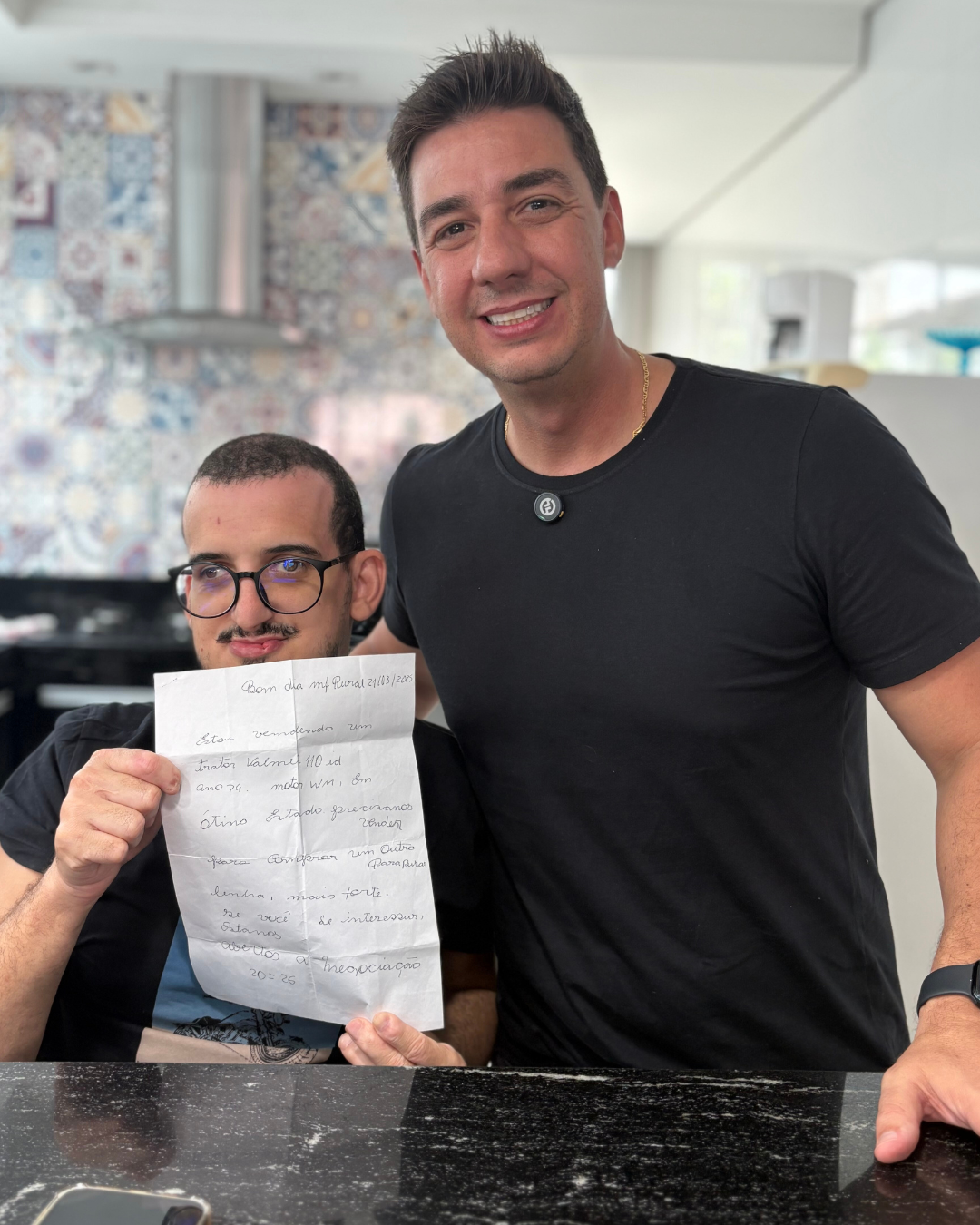 À esquerda, Gustavo, autor da carta, segura o bilhete escrito à mão oferecendo um trator Valmet 110 de 1974. À direita, Rafael Fabrizzi Lucas, CEO do MF Rural, sorri ao lado dele em uma cozinha com azulejos coloridos ao fundo.
