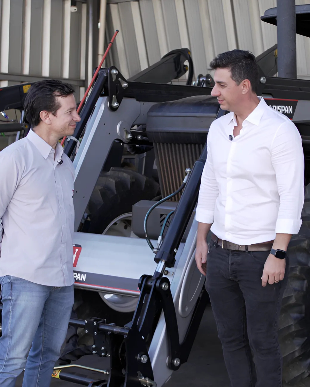 Rafael Fabrizzi Lucas, CEO do MF Rural, conversa com Paulo Nascimento, Diretor da Marispan em frente a um implemento agrícola da Marispan. Ambos estão sorridentes, vestidos de forma social, em um galpão agrícola.