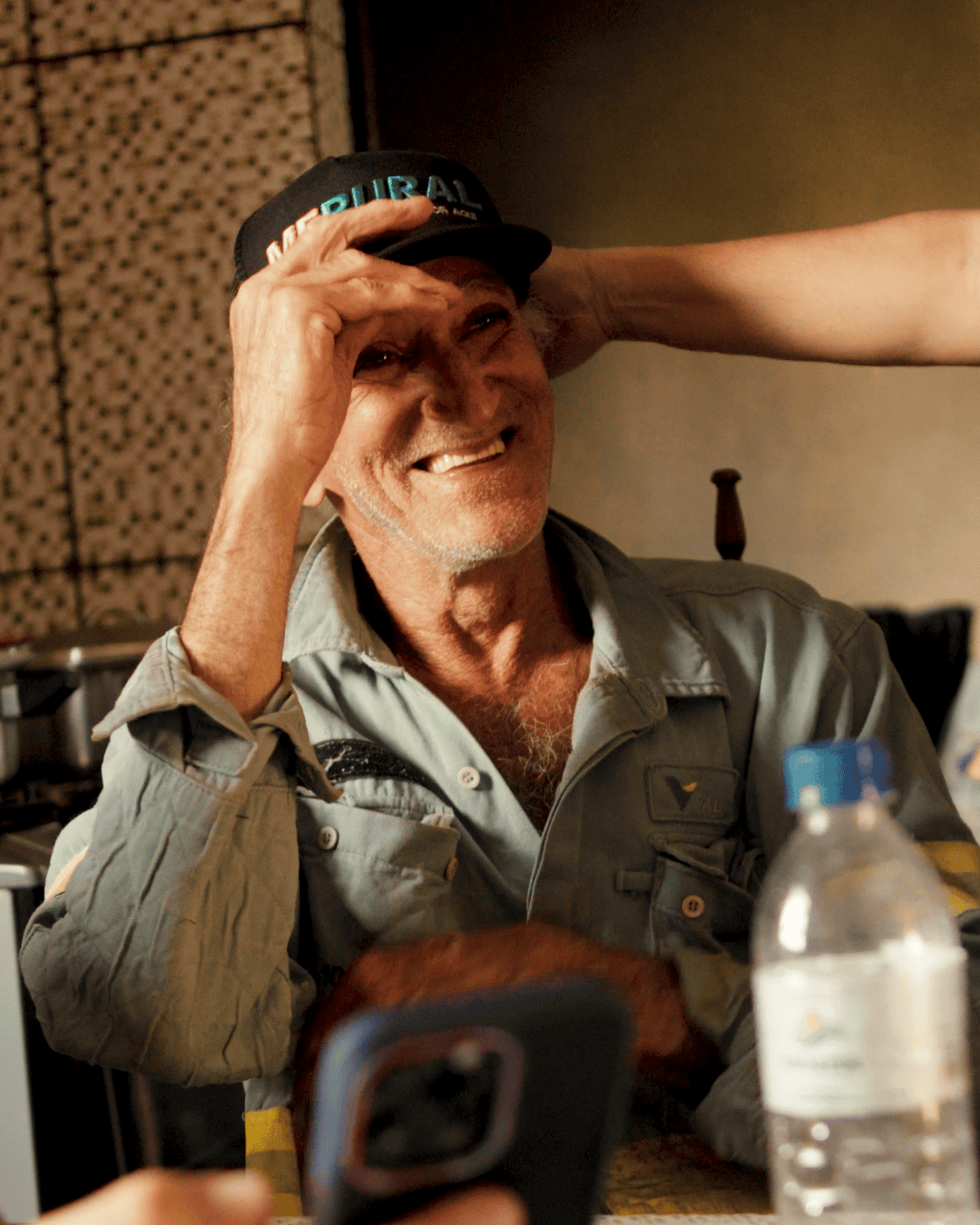 Homem idoso sorri sentado à mesa, usando boné preto com a palavra 'MF Rural' e camisa de trabalho azul clara. Ele segura o boné com uma das mãos enquanto outra pessoa toca sua cabeça de forma carinhosa.