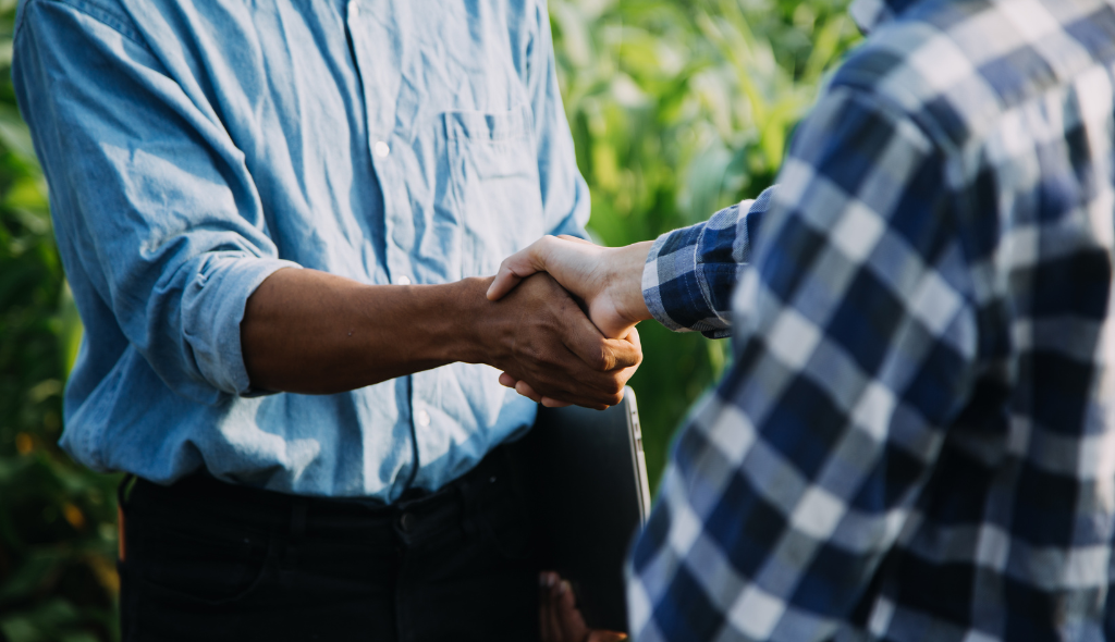 Dois produtores rurais apertando as mãos em acordo no campo, representando contratos de arrendamento e parceria rural.