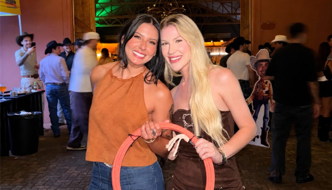Nicoli Fittipaldi e a cantora Ana Castela sorrindo juntas em um evento do universo country, segurando uma corda de la&ccedil;o, com pessoas usando chap&eacute;us ao fundo