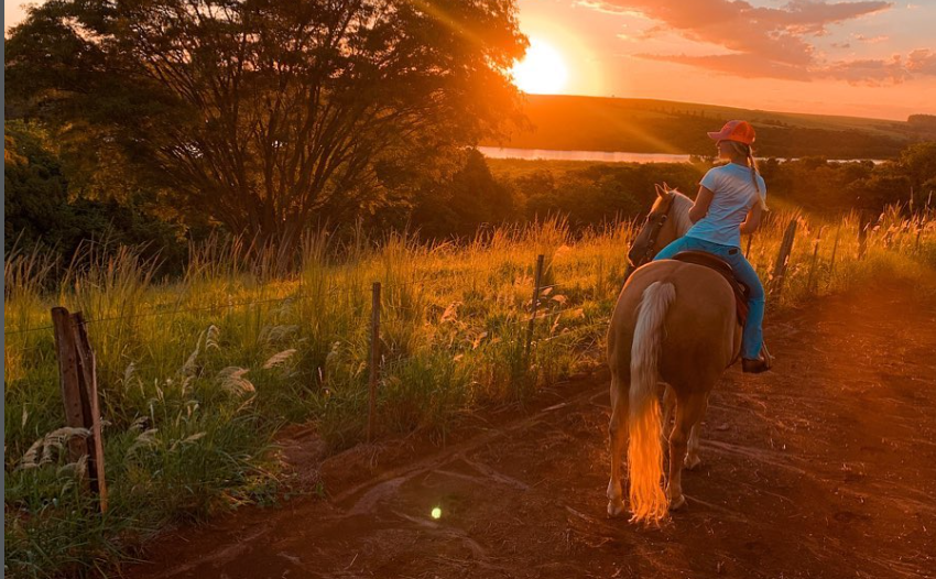 Nicoli de chap&eacute;u rosa e camiseta azul montada em um cavalo, percorrendo uma estrada de terra no campo durante o p&ocirc;r do sol, cercada por &aacute;rvores e vegeta&ccedil;&atilde;o
